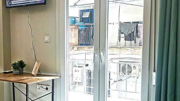 A hotel room with a bed, a TV mounted on the wall, a small table with a plant, and a view through the glass door.