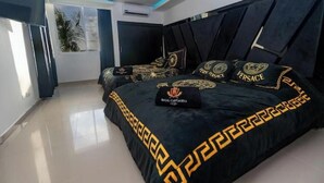 A modern bedroom with a large bed, dark bedding with gold patterns, a black headboard, and a small black pillow with a logo. - Tropical One-Bed Oasis (CARTAGENA BOLIVAR)