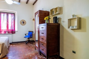 Una stanza con una cassettiera in legno, una sedia, un termosifone, una finestra con tende e una mensola a muro.