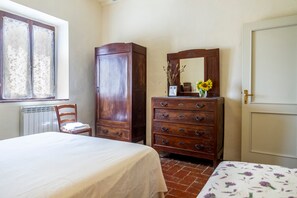 Ein Schlafzimmer mit Bett, Kommode, Stuhl, einem Fenster mit Spitzenvorhängen und einem Heizkörper.