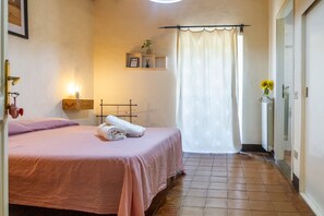 Een slaapkamer met een bed, een nachtkastje, een wandplank met een plant, een raam met gordijn en een betegelde vloer.