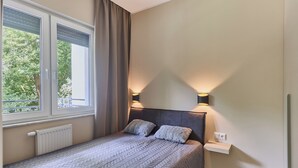 A bedroom with a bed, two bedside lamps, a window with curtains, and a radiator. - 11 listopada 70 c (Świnoujście)