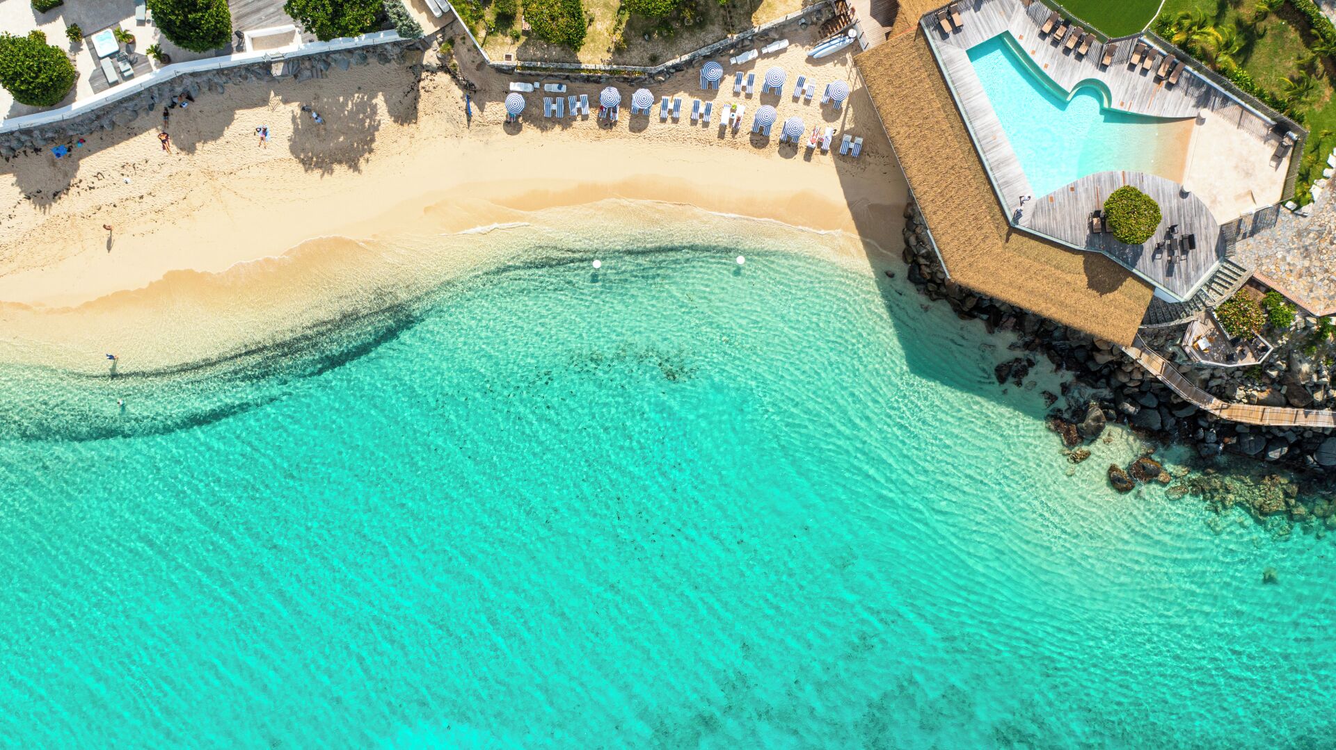 Vue aérienne d’un complexe hôtelier en bord de mer, avec une piscine à l’eau turquoise et limpide, une plage de sable et une terrasse en bois dotée de chaises longues.