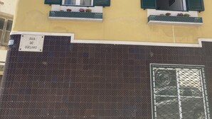 Un bâtiment doté de deux fenêtres, chacune ornée de volets verts, et d’une enseigne indiquant « Rua do Quelhas ».
