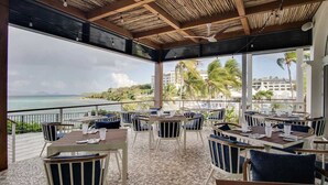 Un espacio para comer al aire libre con mesas y sillas, con vista a una playa y palmeras.