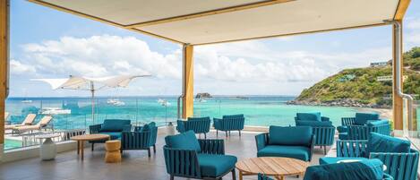 A covered outdoor seating area with chairs and tables, overlooking a body of water with boats and a distant island.