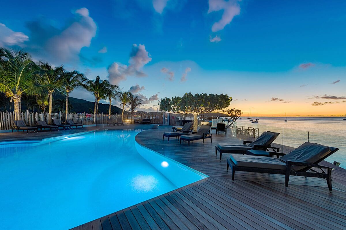 Zona junto a la piscina con sillones de descanso, una terraza de madera y vista al mar al atardecer.