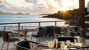 Une terrasse avec des tables et des chaises, donnant sur une étendue d’eau et une masse terrestre lointaine, avec un bâtiment en arrière-plan.