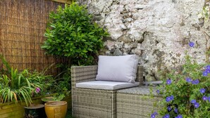 A wicker sofa with a cushion against a stone wall, surrounded by potted plants and a grassy area. - Pass the Keys |Bright Town-Centre Flat with Outdoor Area (Ulverston)