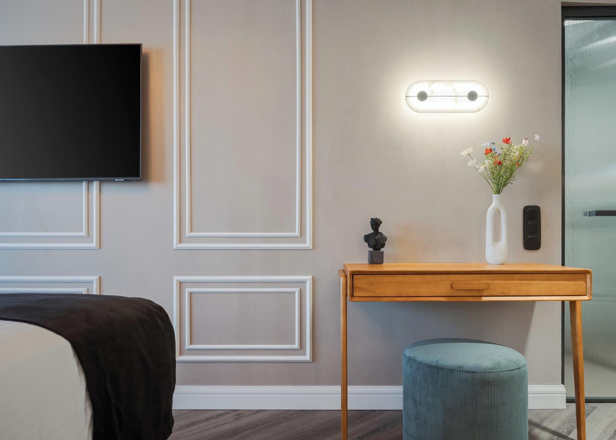 Een hotelkamer met een bed, een aan de muur bevestigde televisie, een nachtkastje met een bloevas en een kleine sculptuur.