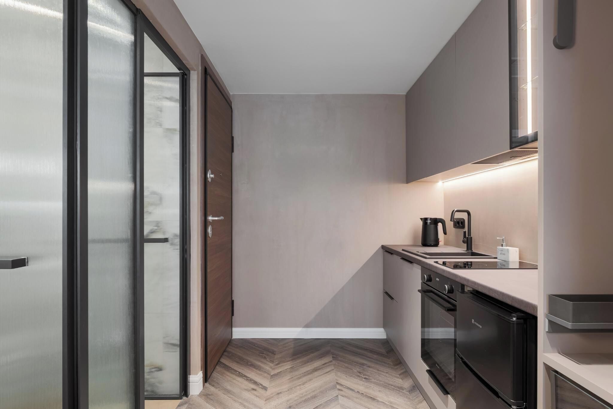 A modern kitchen with a herringbone wood floor, a built-in oven, a stove, and a sink with a kettle.