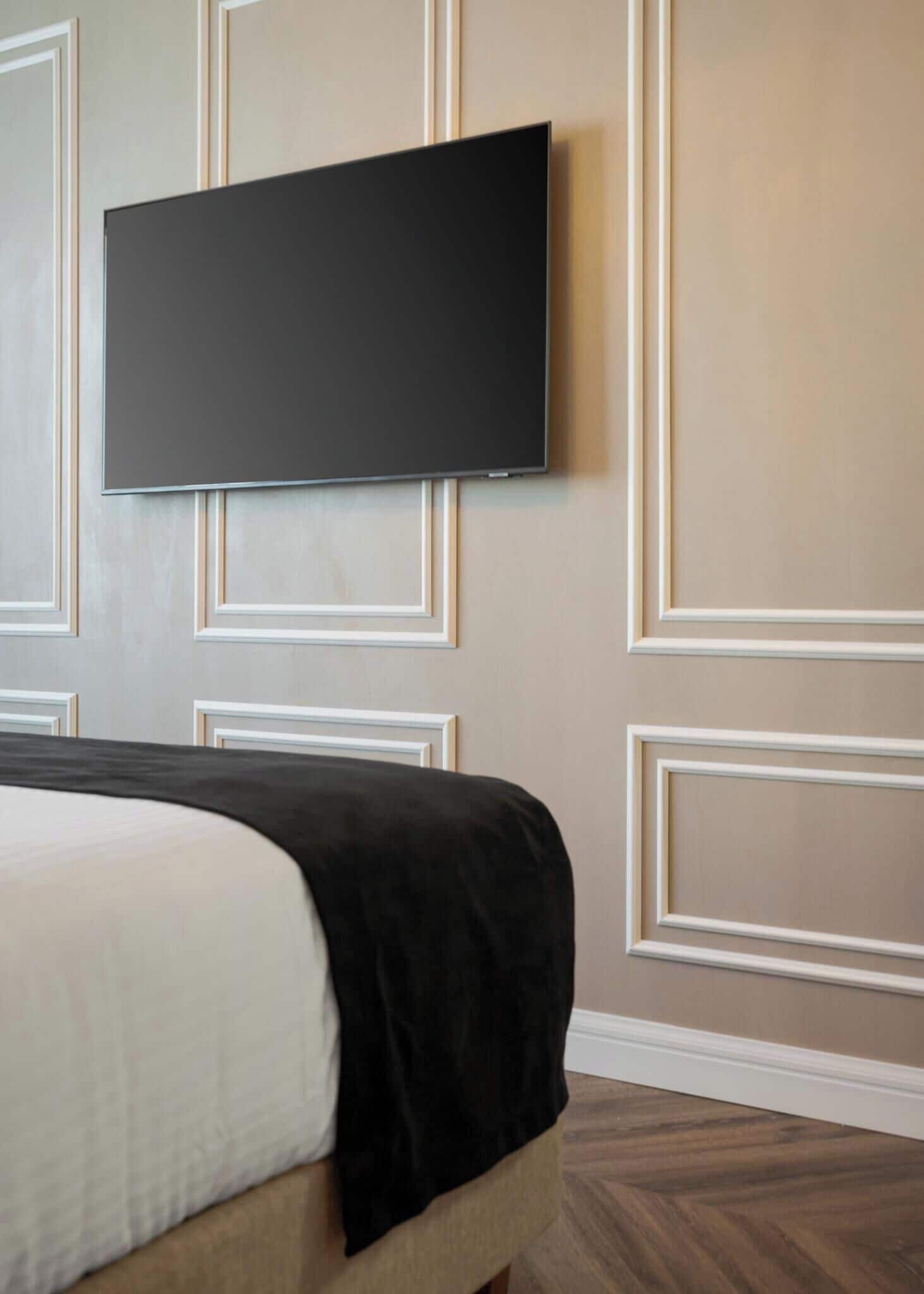 A bedroom with a bed, a flat-screen TV mounted on the wall, and a wooden floor.