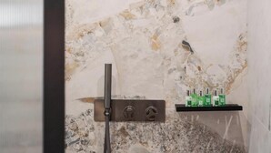 A modern bathroom with a marble wall, a showerhead, and a shelf with toiletries. - Acropolis Luxury Athens Suites (Athens)