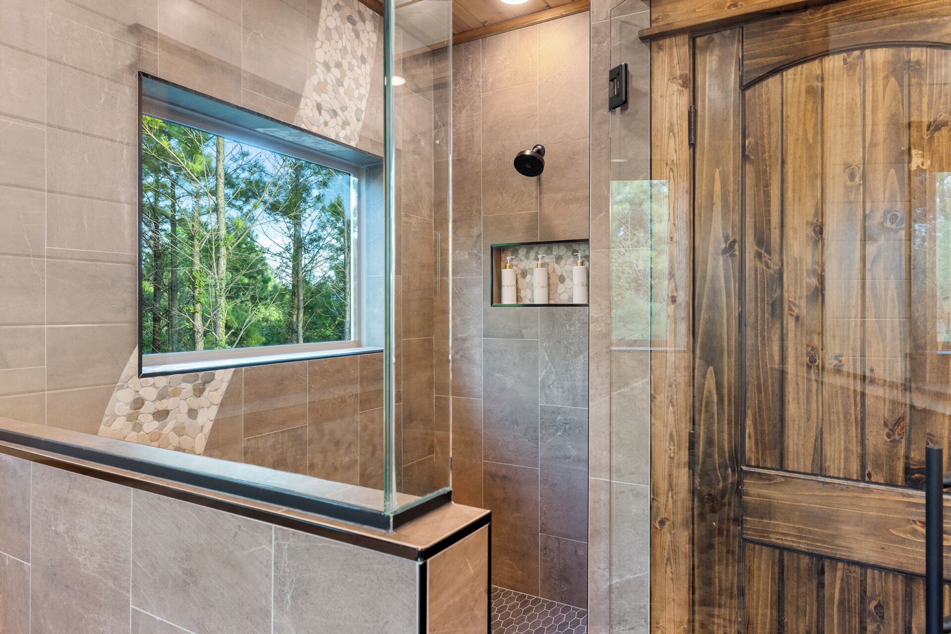 A modern bathroom with a large window, a wooden door, and a built-in shelf with toiletries.