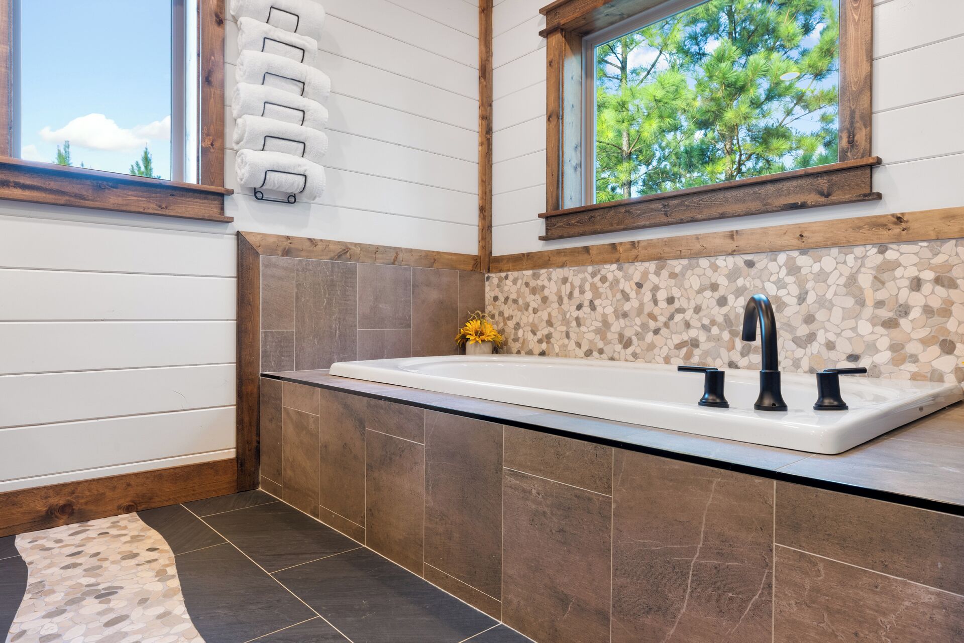 Ein modernes Badezimmer mit einer großen Badewanne, einem Holzfensterrahmen und einer Spritzschutzeinrichtung mit Kieselsteinen.