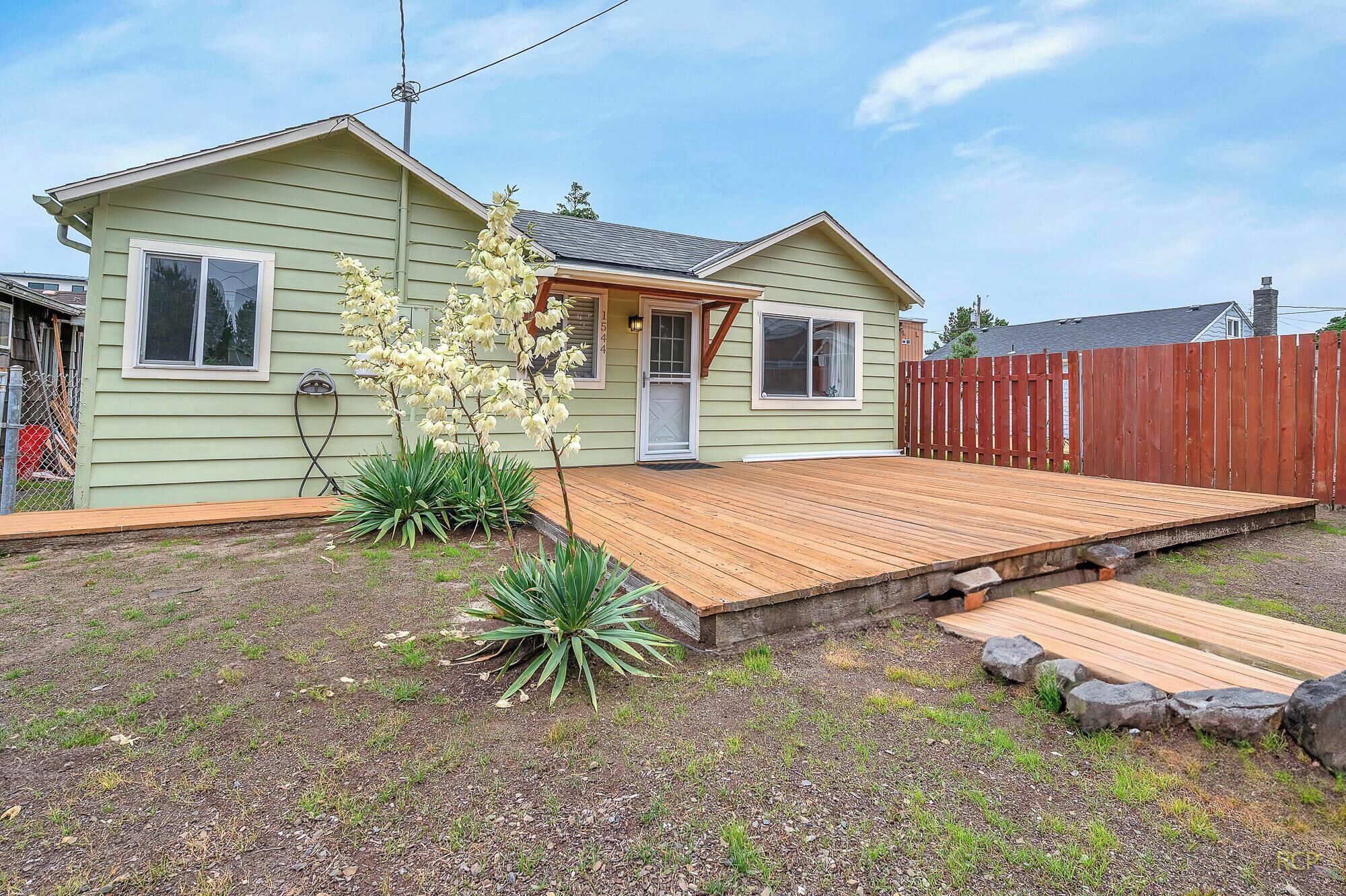 Ein eingeschossiges Haus mit einer Veranda, einer Holzterrasse und einem eingezäunten Garten.