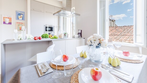 A modern kitchen with a dining area, featuring a large window with a view of a tiled roof and a clear sky. - The White Lotus Penthouse (Lucca)