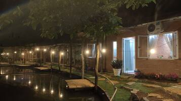 Vue nocturne d’un espace piscine avec des terrasses en bois, un bâtiment en briques dont la porte est ouverte, et des guirlandes lumineuses.