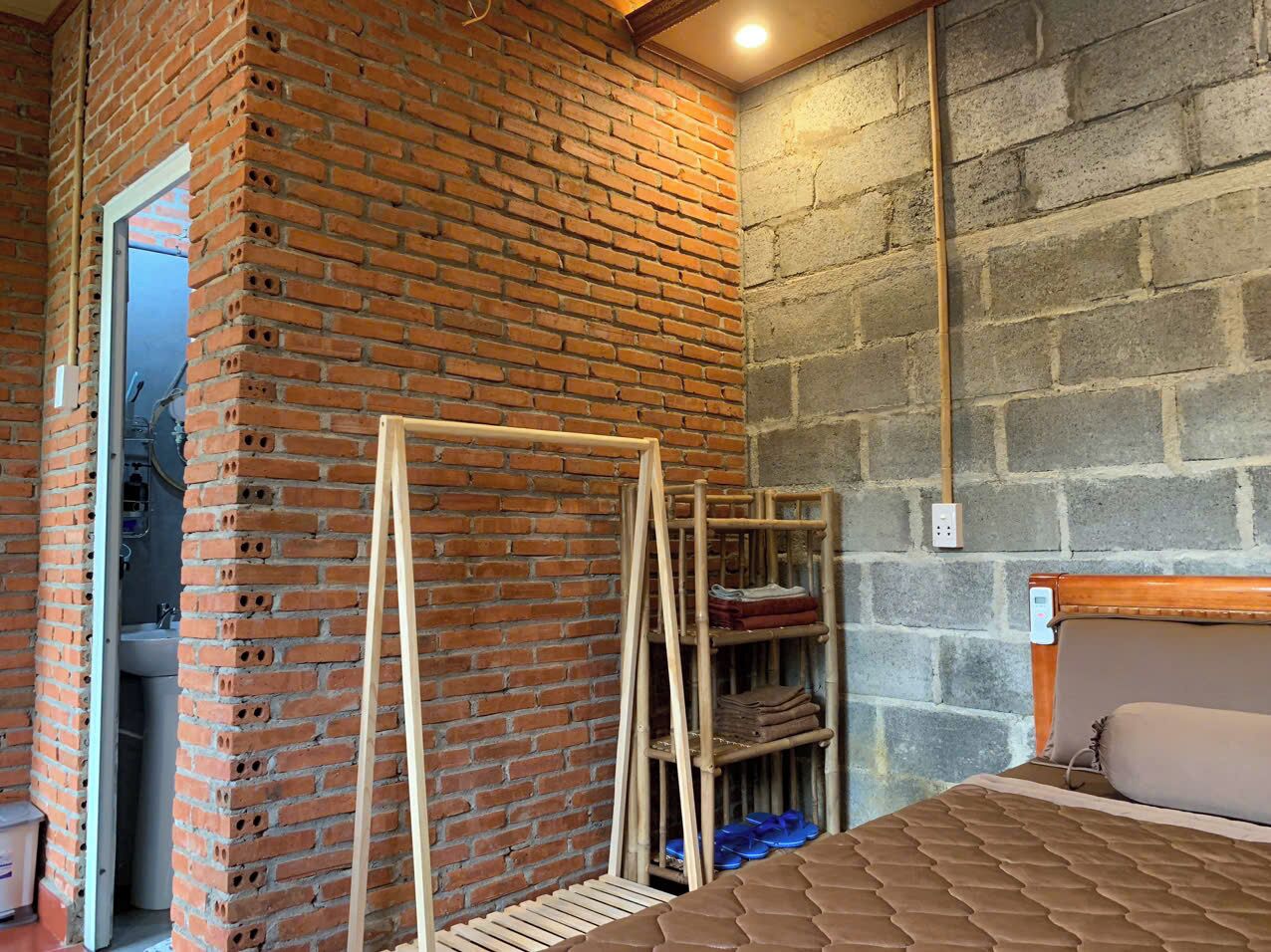 A room with a brick wall, a wooden bed frame, a wooden shelf unit, and a small bathroom visible through a doorway.