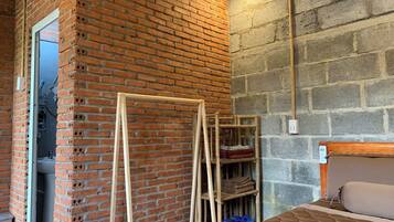 A room with a brick wall, a wooden bed frame, a wooden shelf unit, and a small bathroom visible through a doorway.