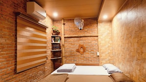A room with a brick wall, a wooden shelf with books and bottles, a bed with a brown blanket, and a fan on the ceiling.