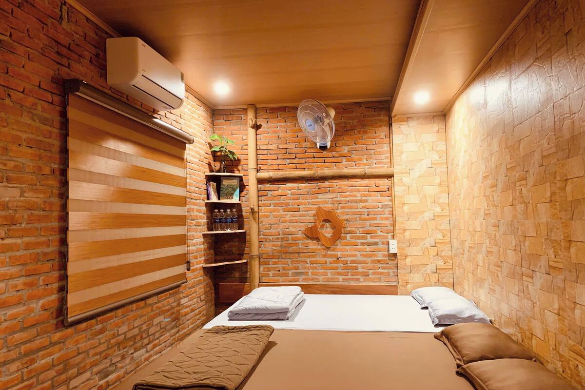 Una habitación con pared de ladrillo, una estantería de madera con libros y botellas, una cama con manta marrón y un ventilador en el techo.