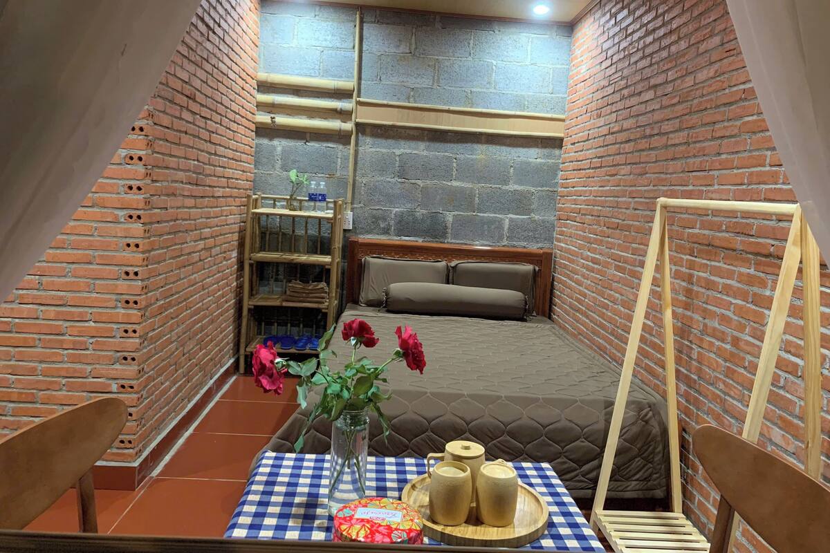 Una habitación acogedora con cama, una mesa con mantel a cuadros, un jarrón con flores y una estantería con objetos.