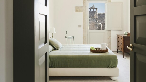 A bedroom with a bed, a dresser, a lamp, and a window with a view of a building. - La Canonica Suites (Cannole)