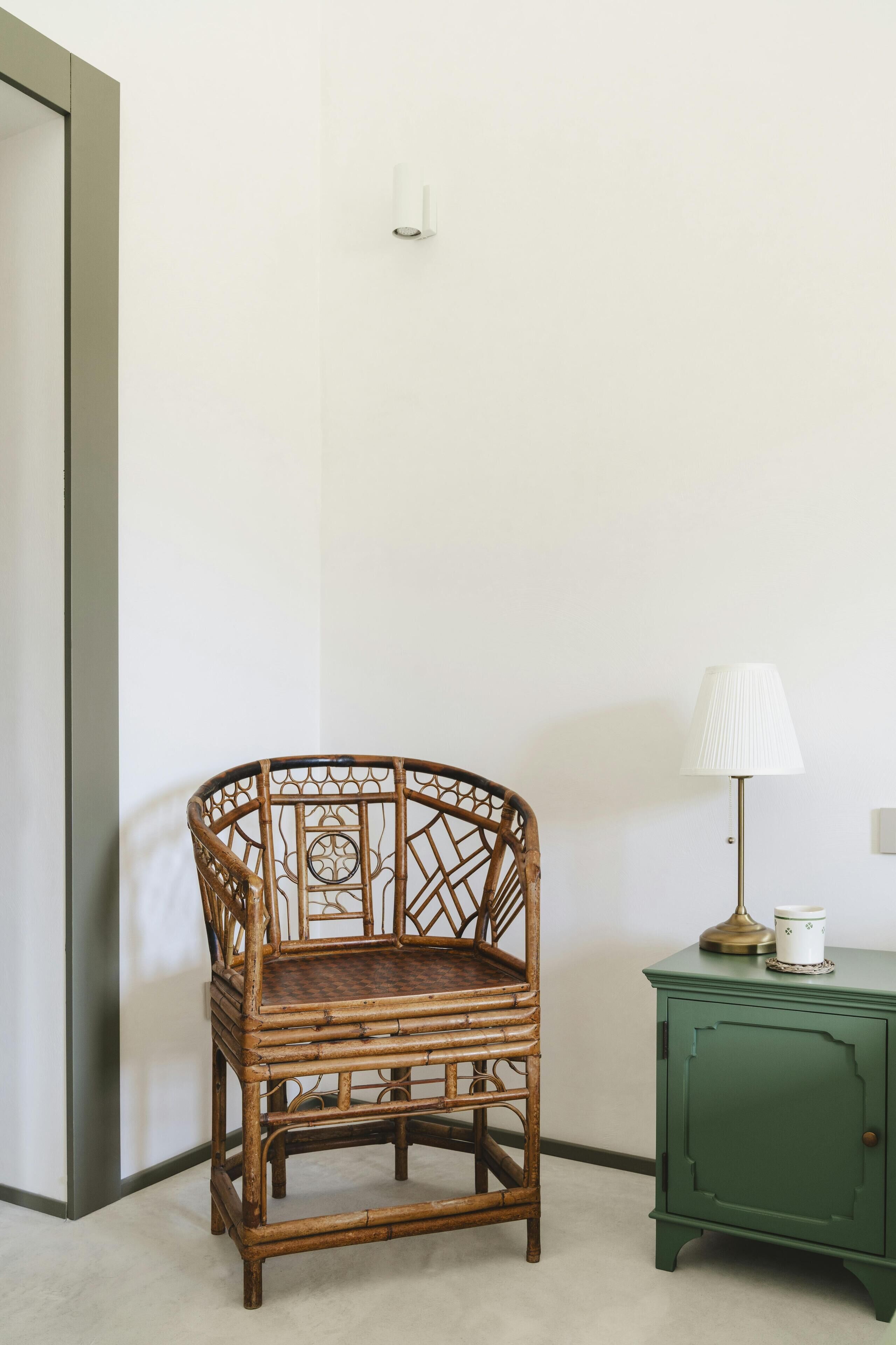 Una silla de mimbre en un rincón de una habitación, junto a un armario verde y una lámpara.
