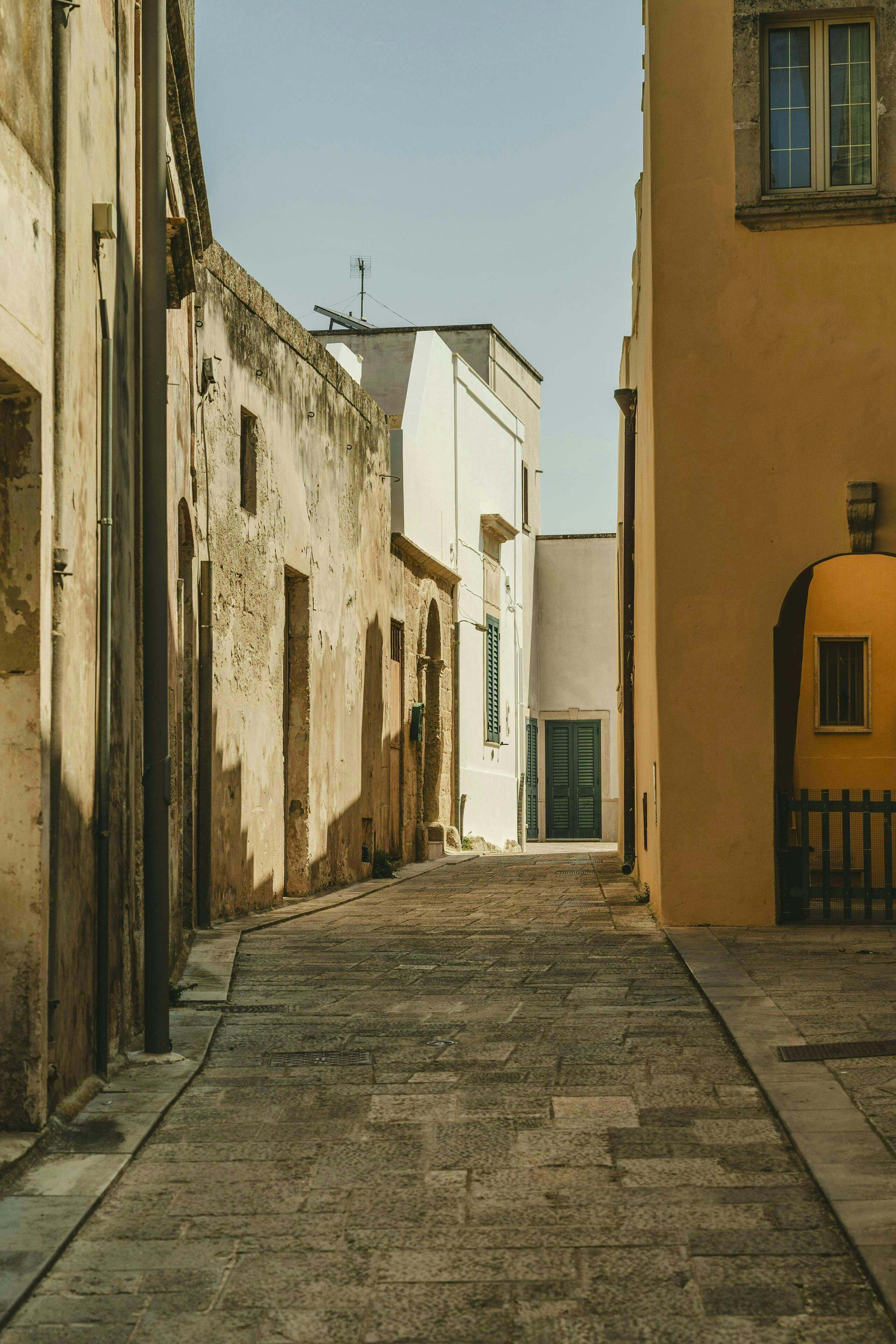 A narrow street with buildings on either side, featuring a mix of architectural styles and a cobblestone pavement.