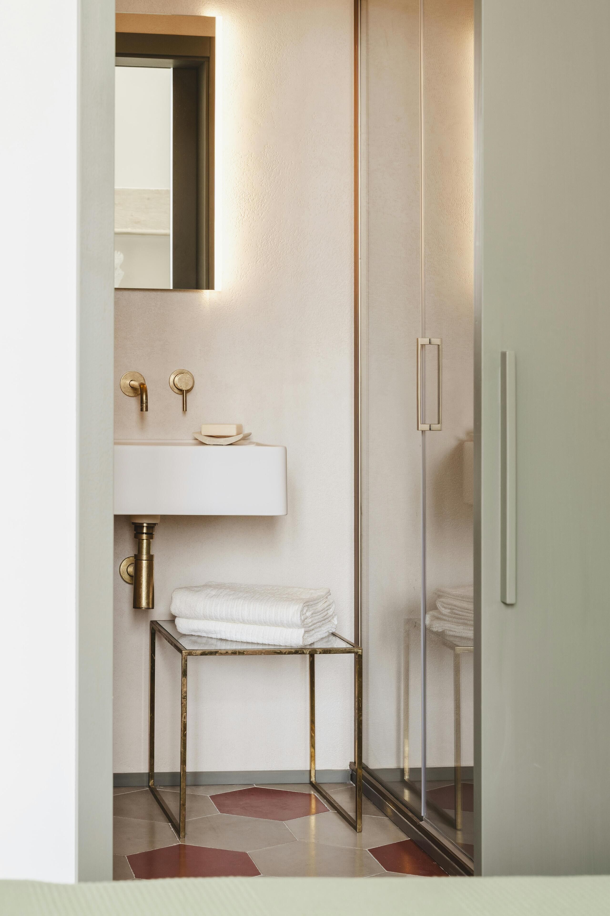 Ein modernes Badezimmer mit einer Duschkabine aus Glas, einem weißen Waschbecken mit goldfarbenen Armaturen und einem kleinen Tisch mit Handtüchern.