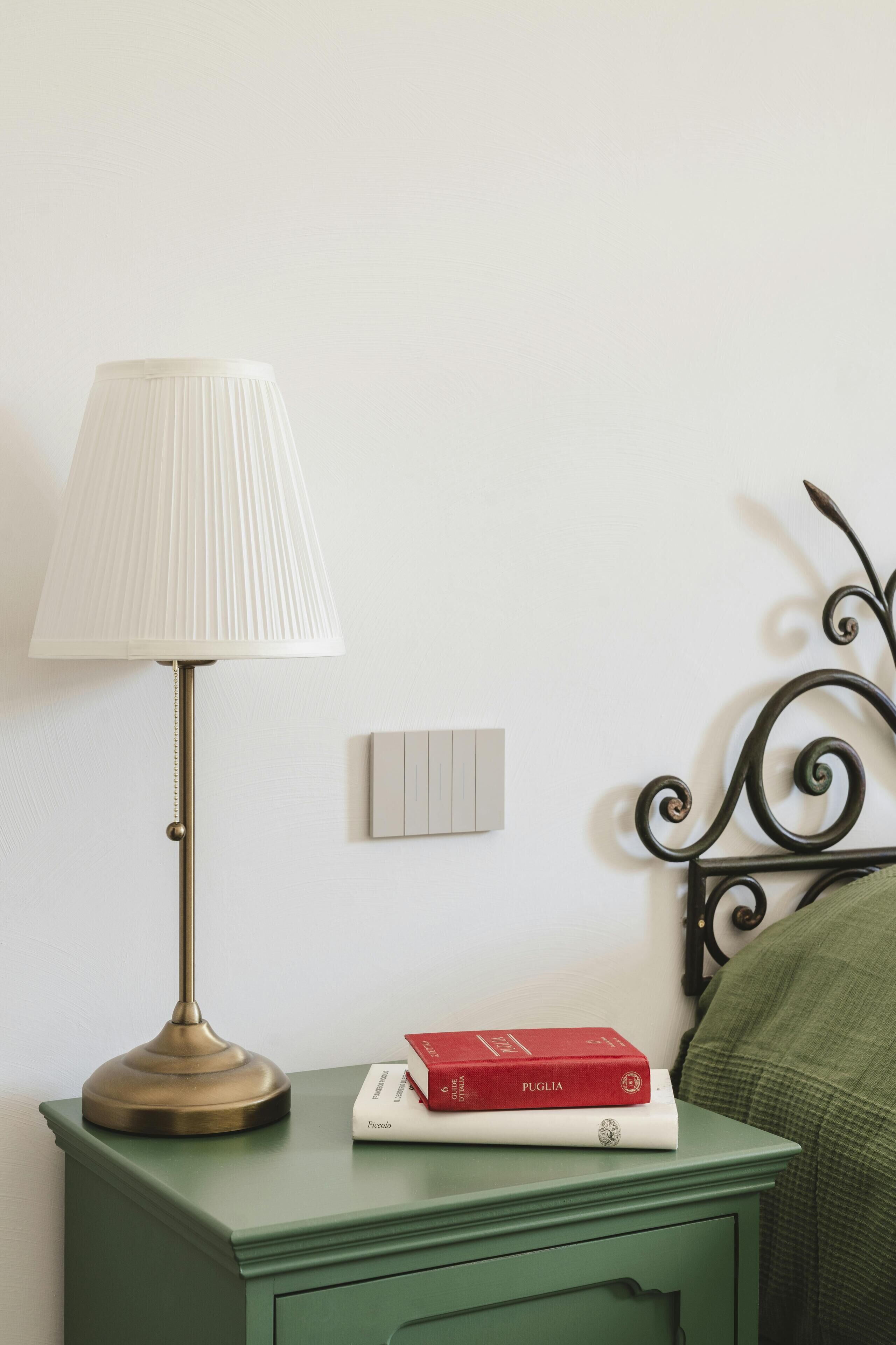 A green bedside table with a white lamp and two books, one red and one white, against a white wall.