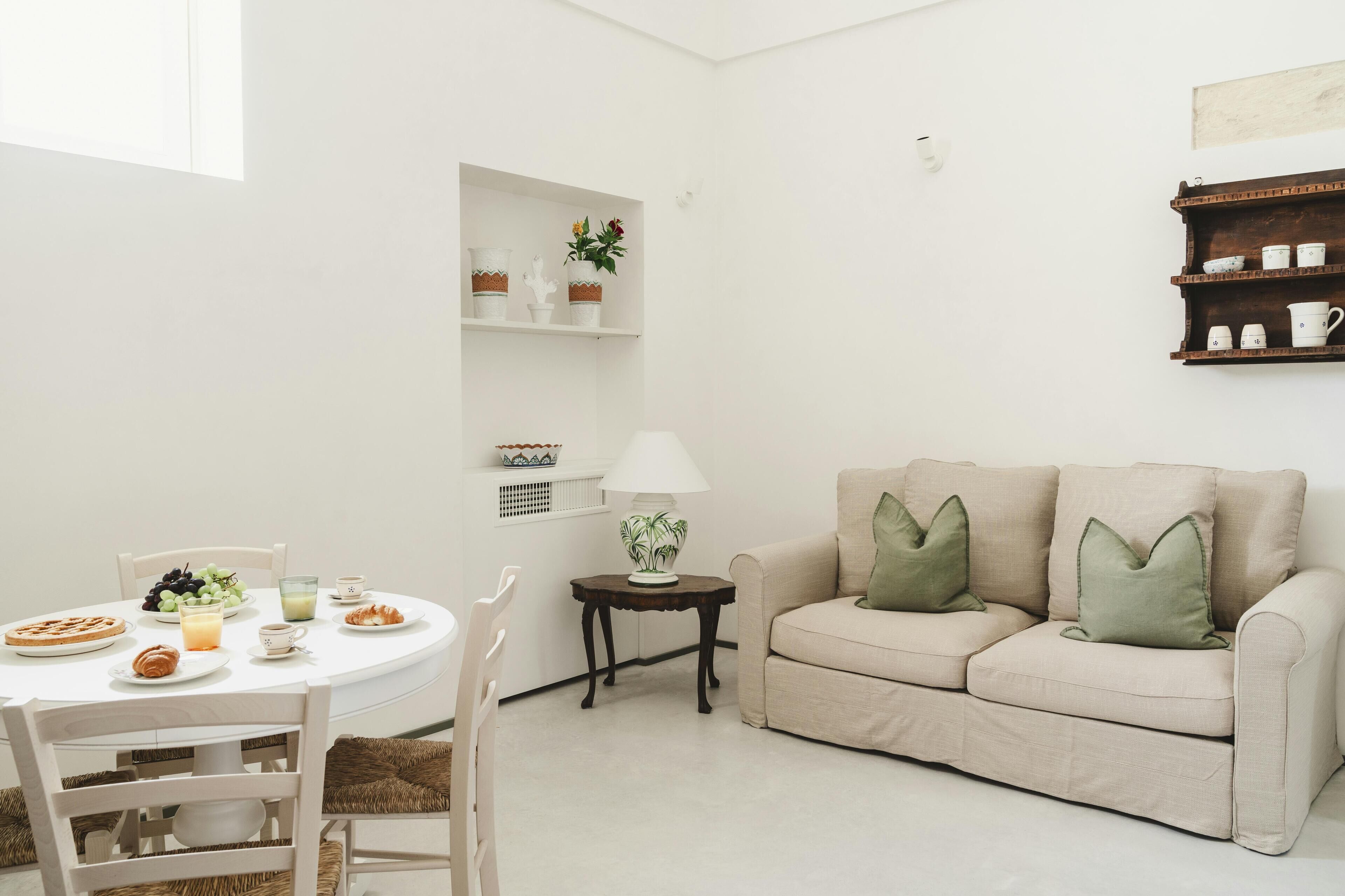 A cozy living room with a round dining table, a beige sofa with green cushions, and a small side table with a lamp.