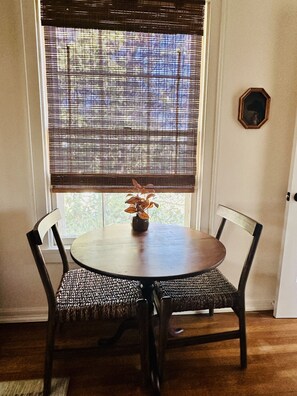 A dining area with a round table, two chairs, a window with blinds, and a small vase with flowers.