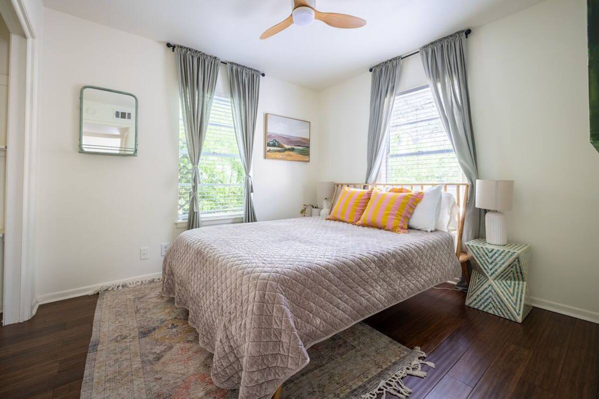 A bedroom with a bed, a ceiling fan, a nightstand, a lamp, a rug, and a window with curtains.
