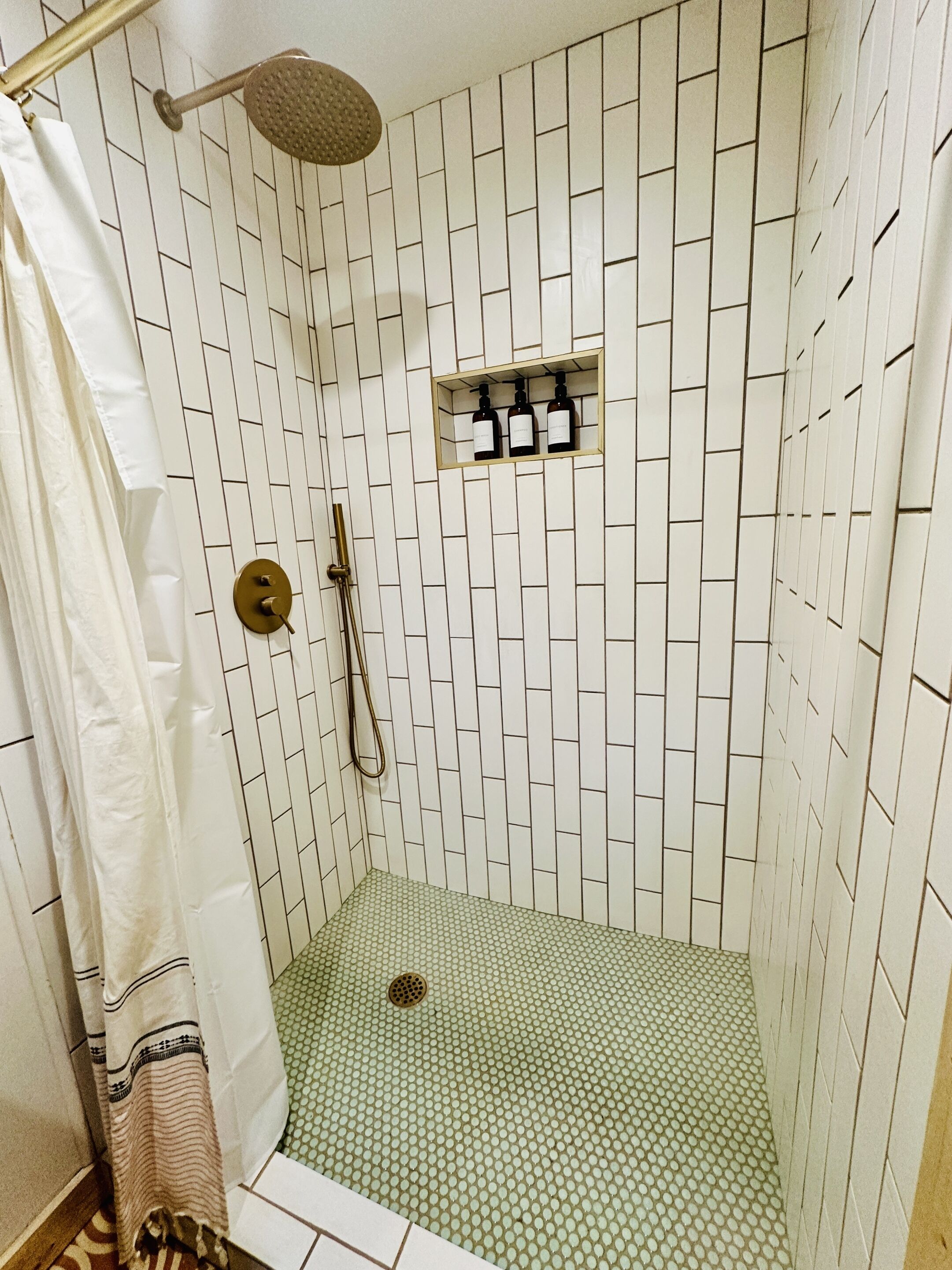 Une douche avec un mur recouvert de carreaux blancs, un pommeau de douche et une petite étagère sur laquelle sont posées des bouteilles.