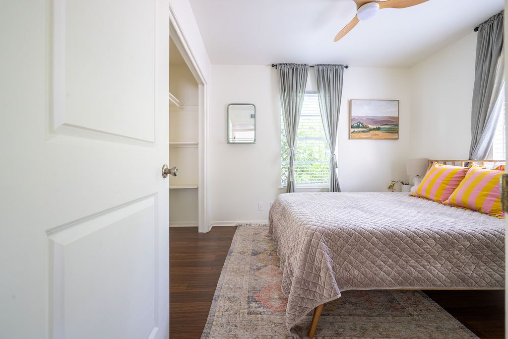 A bedroom with a bed, a ceiling fan, a window with curtains, and a painting on the wall.