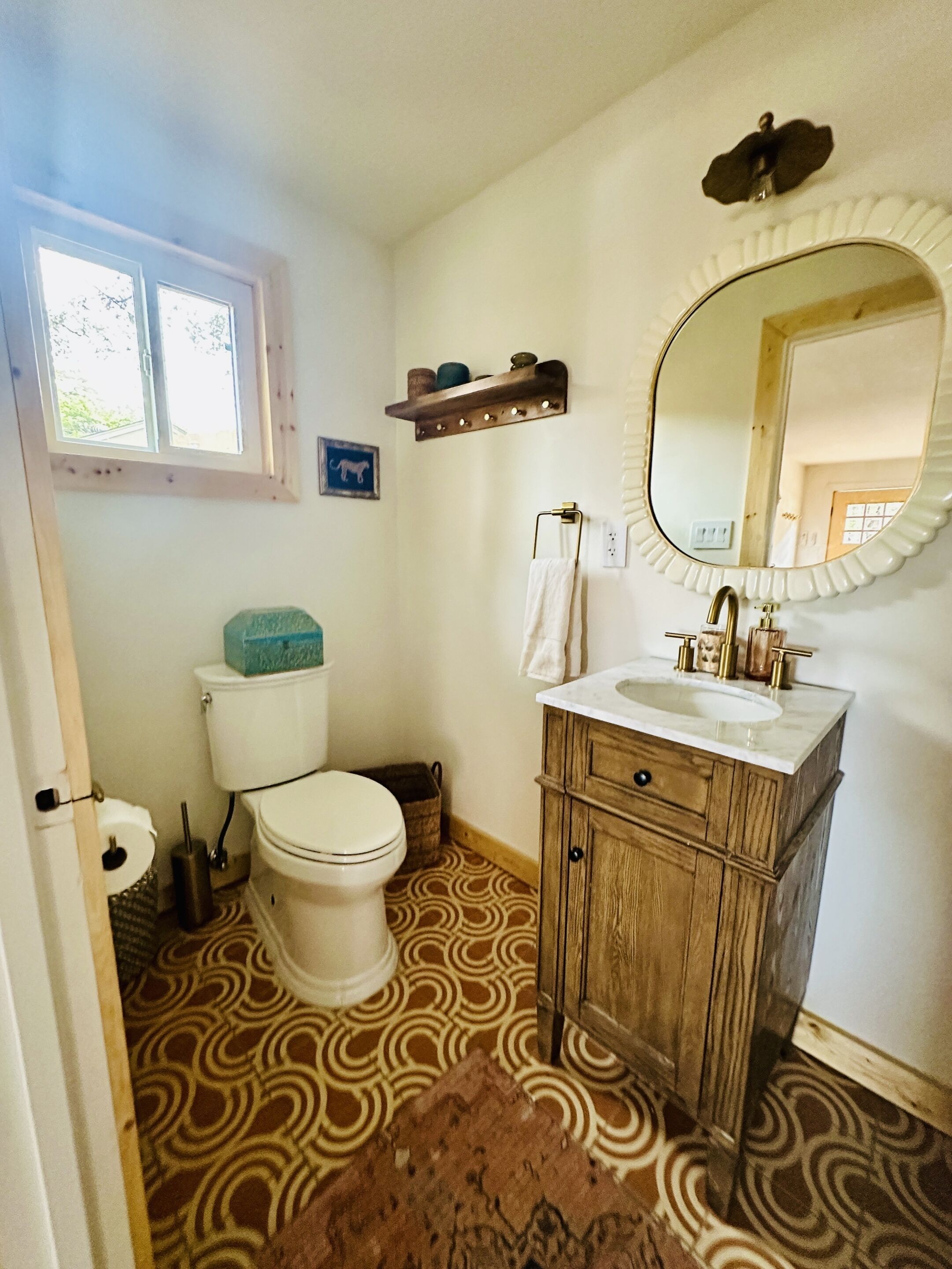 A bathroom with a toilet, sink, and mirror. There is a window above the toilet and a shelf with decorative items above the sink.
