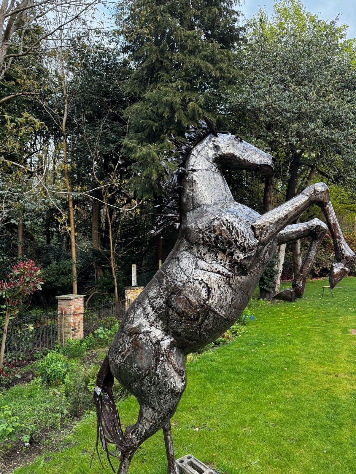 A metal sculpture of a rearing horse in a grassy area with trees and a brick structure in the background.