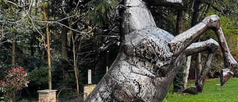 En skulptur av en häst, tillverkad i metall och avbildad i en stående, upprätt position, placerad i ett grönområde med träd och en byggnad av tegel i bakgrunden.