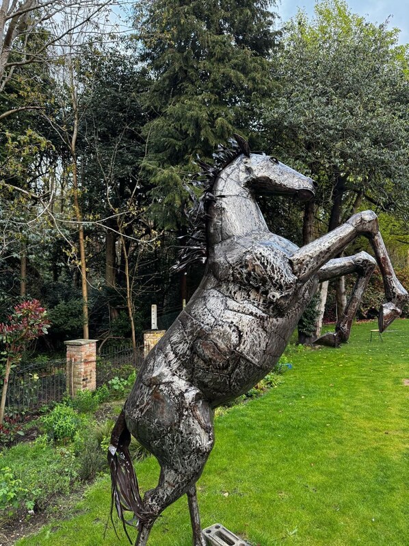 A metal sculpture of a rearing horse in a grassy area with trees and a brick structure in the background.