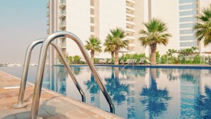 A swimming pool with metal ladders, surrounded by palm trees and lounge chairs, with tall buildings in the background.