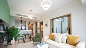 Ein modernes Wohnzimmer mit einem weißen Sofa, einem grünen Sessel und einem Couchtisch. Im Bereich befindet sich eine Essecke mit Tisch und Stühlen, und im Hintergrund ist eine Küche zu sehen.