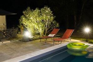 A poolside area with two lounge chairs, a table with chairs, and a small table with a lamp.