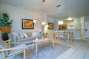 A modern living room with a sofa, armchairs, a coffee table, and a dining area with a table and chairs. There is a kitchen in the background with cabinets and a microwave.