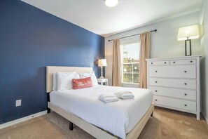 A neatly made bed with white bedding and a red pillow, a white dresser with drawers, a lamp on the dresser, and a window with curtains.
