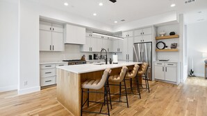 A modern kitchen with white cabinetry, a central island with bar stools, stainless steel appliances, and a wooden floor. - The Eastwood Escape (Dallas)