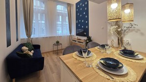 Un coin repas moderne avec une table en bois dressée pour deux personnes, un canapé agrémenté d’un ours en peluche et un téléviseur posé sur un meuble.