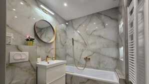 Une salle de bain moderne avec un mur en marbre, un miroir rond, un lavabo blanc, des toilettes et une baignoire.