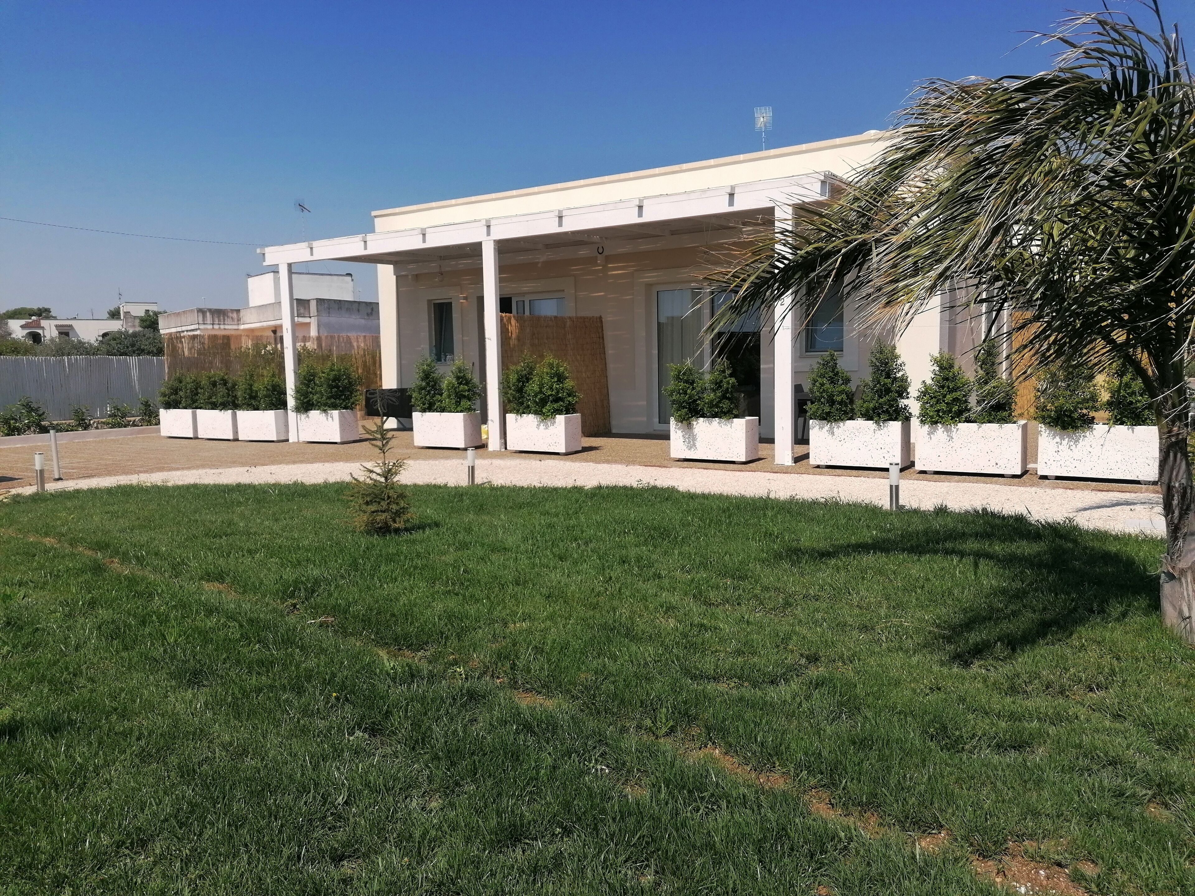 Un edificio moderno con un espacio exterior cubierto, rodeado de césped cuidado y plantas en macetas.